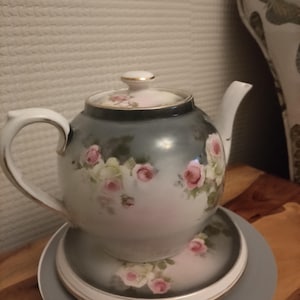 May include: A white porcelain teapot with a floral design featuring pink and white roses. The teapot has a gold trim and sits on a matching saucer.
