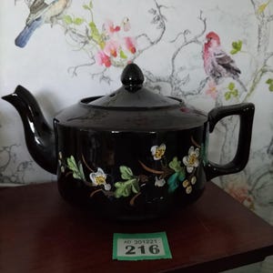 May include: Black teapot with a floral design featuring white flowers, green leaves, and gold accents. The teapot is sitting on a wooden surface with a green price tag that reads "AD 301221 216".