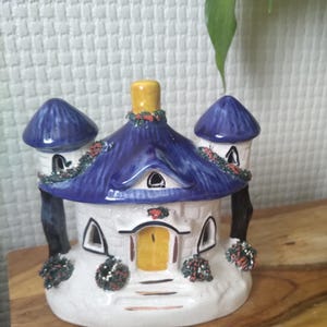 May include: A ceramic house figurine featuring a white stone-textured base, a bright blue roof with two side turrets, and a yellow door. The structure is decorated with small painted green and red wreaths and has a yellow chimney on top.