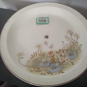 Può includere: Un piatto rotondo in ceramica color bianco sporco con un delicato motivo floreale nei toni del blu, verde e marrone. Il bordo presenta una sottile trama a coste e un profilo metallico. Un piccolo adesivo rettangolare verde riporta il testo 560.