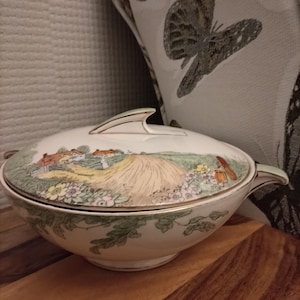 Könnte beinhalten: Eine weiße Keramik-Servierschüssel mit Deckel. Die Schüssel hat einen grünen und goldenen Rand und eine bemalte Szene einer Landschaft mit einem Bauernhaus und einem Heufeld.