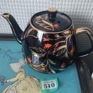 Peut inclure: Théière en céramique noire avec garniture dorée et motif floral. La théière a un couvercle avec un bouton doré et un bec verseur incurvé. Le motif floral comprend des accents dorés et orange. La théière est posée sur un plateau bleu.