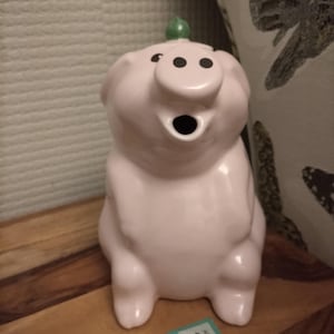 以下が含まれることがあります： 驚いた表情のピンク色のセラミック豚の置物。頭には小さな緑色のアクセントがあります。豚は黒い目をし、口を開けています。置物は木製の表面に立っています。
