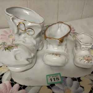 以下が含まれることがあります： 金色の縁取りと花柄が施された、白い磁器製のブーツ型のプランター3個セット。一番大きなブーツは他の2つよりも背が高いです。