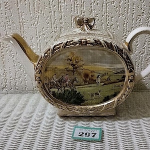 Op de afbeelding: Crèmekleurige keramische theepot met een goudomrande ovale landschapsscène. De scène toont figuren te paard, honden en een landelijk landschap. De handgreep en tuit zijn wit met gouden accenten. Het deksel heeft een decoratieve strik.
