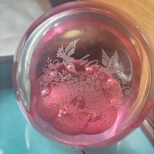 Può includere: Un fermacarte in vetro trasparente con interno rosa. L'interno rosa presenta un motivo floreale e due uccelli incisi. Il fermacarte è rotondo e poggia su una superficie blu.