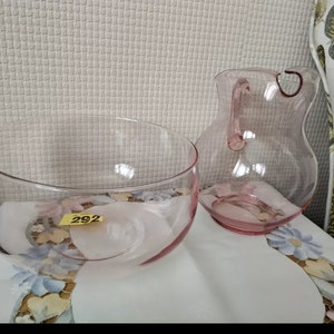 May include: A set of two pink glass serving pieces, a bowl and a pitcher. The bowl is round and has a wide rim. The pitcher has a handle and a rounded body. Both pieces are clear glass with a pink tint.