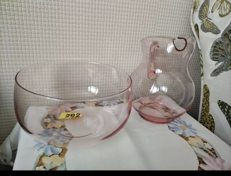 May include: A set of two pink glass serving pieces, a bowl and a pitcher. The bowl is round and has a wide rim. The pitcher has a handle and a rounded body. Both pieces are clear glass with a pink tint.