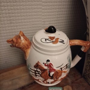 Pode incluir: Uma chaleira de cerâmica branca com um bico e uma alça em forma de cabeça de raposa. A chaleira apresenta uma cena de caça com um cavaleiro a cavalo. A tampa tem um botão preto e detalhes de raposa. Um item decorativo.