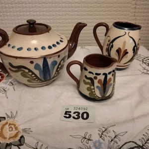 Può includere: Un set di tre pezzi in ceramica per il tè, in base bianca con disegni dipinti in marrone, verde, blu e arancione. Il set comprende una teiera, un creamer e uno zuccheriera.
