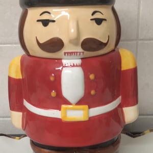 Puede incluir: Un tarro de galletas de cerámica con forma de cascanueces, con un sombrero negro alto, chaqueta de uniforme roja con detalles amarillos, corbatín blanco y botas marrones. La figura tiene bigote y cejas pintados. Una etiqueta verde muestra AD 301221 531.