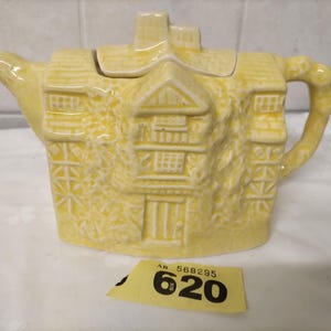 May include: A yellow ceramic teapot shaped like a house, featuring detailed architectural elements such as windows, doors, and a roof. The teapot has a handle and a spout. A yellow tag with the number "620" is in the foreground.