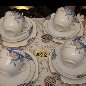 May include: Four sets of vintage teacups and saucers with floral designs. Each set includes a cup, saucer, and a larger plate. The cups and saucers are white with blue and pink floral patterns. The plates are white with blue and pink floral borders.