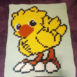 May include: A crocheted blanket featuring a yellow, pixelated chick with red and black accents. The chick is standing on white clouds with black outlines. The blanket is on a purple surface.