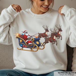 Puede incluir: Sudadera blanca con un gráfico festivo de Papá Noel en un trineo azul tirado por tres renos. Papá Noel lleva un traje y un gorro rojos. El trineo tiene detalles dorados y luces intermitentes.