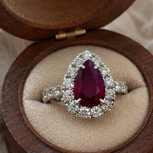 May include: A pear-shaped ruby ring with a halo of diamonds, set in a silver band. The ring is displayed in an open, wooden jewelry box with a beige interior. The ruby is a deep red color.