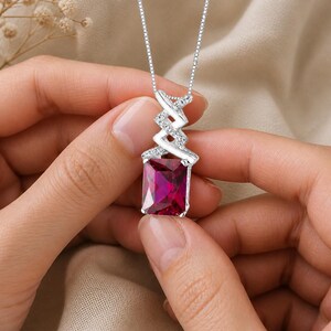 May include: A silver necklace with a rectangular, deep red gemstone pendant. The pendant is set in a silver frame and hangs from a delicate chain. The necklace is held by a person's hands. The text "MADE TO SHINE" is at the bottom.