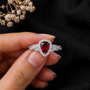 Puede incluir: Un anillo plateado con una piedra preciosa roja oscura en forma de pera en el centro, rodeada por un halo de piedras claras más pequeñas. La banda del anillo presenta piedras claras adicionales, creando un efecto brillante. Fondo negro.
