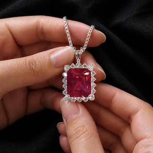 May include: A silver necklace with a large, rectangular, ruby-colored gemstone pendant. The pendant is surrounded by smaller, clear gemstones. The necklace is held against a black background. The text "MADE TO SHINE" is at the bottom.