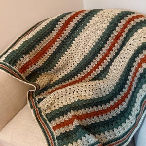Puede incluir: Una manta de ganchillo hecha a mano con un patrón de rayas en tonos verdes, óxido y crema. La manta está doblada sobre una superficie de color claro, mostrando las puntadas texturizadas y las variaciones de color. Los bordes tienen un borde contrastante.