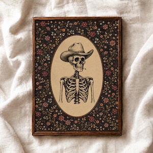 Può includere: Un'opera d'arte da parete rettangolare incorniciata con un'illustrazione ovale centrale di uno scheletro che indossa un cappello da cowboy e tiene una sigaretta. Lo sfondo è un motivo floreale scuro con piccoli fiori rosa, arancioni e gialli su nero.