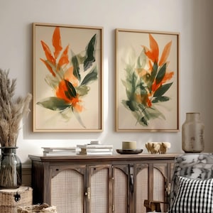 Könnte beinhalten: Zwei gerahmte abstrakte Kunstdrucke. Jeder Druck zeigt ein Aquarelldesign aus orangefarbenen und grünen Blättern auf cremefarbenem Hintergrund. Die Rahmen sind hellbraun. Das Kunstwerk wird über einem Holzschrank mit gewebten Korbtüren ausgestellt.