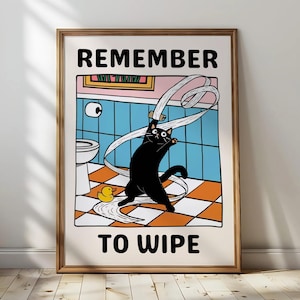 Op de afbeelding: Ingelijste poster met een cartoon zwarte kat die speelt met toiletpapier in een badkamer. De tekst "REMEMBER TO WIPE" staat in het zwart. Een gele badeend ligt op de geblokte vloer.
