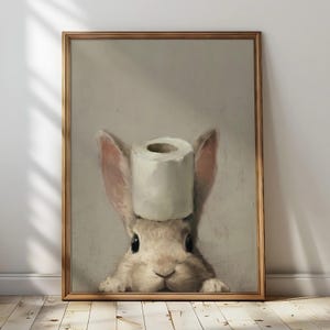 Puede incluir: Impresión enmarcada que presenta un conejo con un rollo de papel higiénico en la cabeza. La obra de arte tiene un estilo vintage con colores suaves y un fondo neutro. El marco es de madera natural cálida.