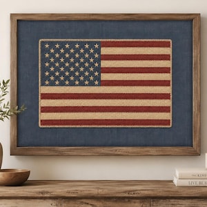 May include: A framed wall art piece featuring a textured, stitched American flag with a blue canton of stars and alternating red and cream-colored stripes. The flag is mounted on a dark blue fabric background within a rustic brown wooden frame.