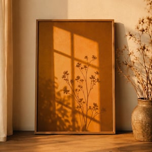 Könnte beinhalten: Ein gerahmter botanischer Druck mit warmem, orangefarbenem Hintergrund. Der Druck zeigt eine Silhouette zarter floraler Stiele und Blätter. Der Rahmen ist hellbraun und das Bild vor einer Wand mit einem Fensterschatten.