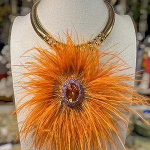 May include: A gold-toned metal collar necklace featuring a large, circular arrangement of vibrant orange feathers. At the center of the feathers is an oval-shaped orange gemstone surrounded by a border of small, sparkling purple-toned stones.