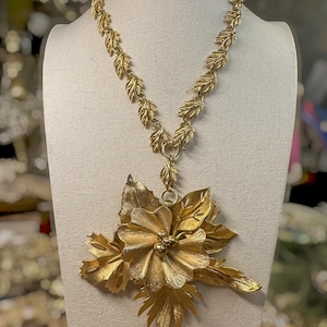 May include: A gold-toned necklace featuring a chain composed of interconnected metallic leaves. The centerpiece is a large, dimensional floral pendant with layered petals and foliage, accented by small spherical beads at the center of the flower.