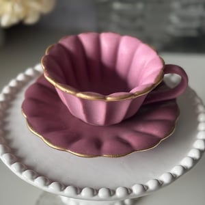 May include: A pink teacup and saucer set with a gold trim, resting on a white cake stand. The teacup has a scalloped edge and a matching saucer. The cake stand has a beaded edge.