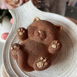 Puede incluir: Un postre con forma de oso marrón con detalles dorados, sobre un plato de cerámica blanca moteada. El oso tiene detalles dorados en las orejas y las patas. El plato tiene una forma orgánica.