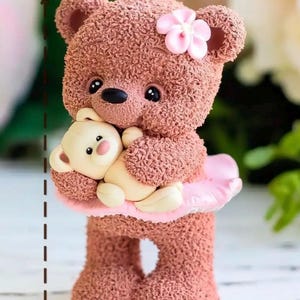 May include: A handcrafted, brown teddy bear figurine, 7.5 cm tall and 4.5 cm wide, holding a smaller cream-colored bear. The bear wears a pink tutu and a pink flower on its head. The figurine weighs approximately 55 grams.