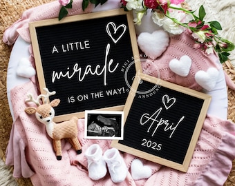 May include: A black letter board with the text "A little miracle is on the way!" and a heart. A second black letter board with the text "April 2025" and a heart. A pink blanket with a deer toy, baby booties, and white hearts.