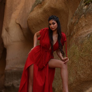 Puede incluir: Una mujer con un vestido rojo maxi con escote pronunciado y aberturas laterales se encuentra en un ca&ntilde;&oacute;n rocoso. El vestido tiene un lazo de cuerda en la cintura y la mujer lleva el pelo largo y oscuro suelto.