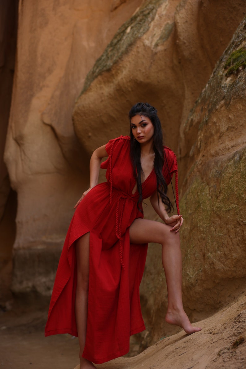 Puede incluir: Una mujer con un vestido rojo maxi con escote pronunciado y aberturas laterales se encuentra en un ca&ntilde;&oacute;n rocoso. El vestido tiene un lazo de cuerda en la cintura y la mujer lleva el pelo largo y oscuro suelto.