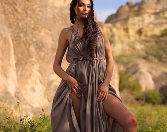 Puede incluir: Una mujer con un vestido largo, fluido y gris se encuentra en un campo de flores amarillas. Tiene el pelo largo y oscuro y mira hacia un lado. El fondo es un paisaje rocoso.
