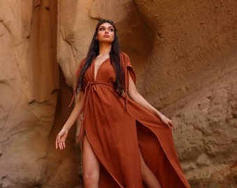 May include: A woman wearing a long, rust-colored dress with a deep V-neckline and side slits. The dress has a tie at the waist and is made of a lightweight fabric. The woman is standing in a desert landscape with rocky cliffs in the background.