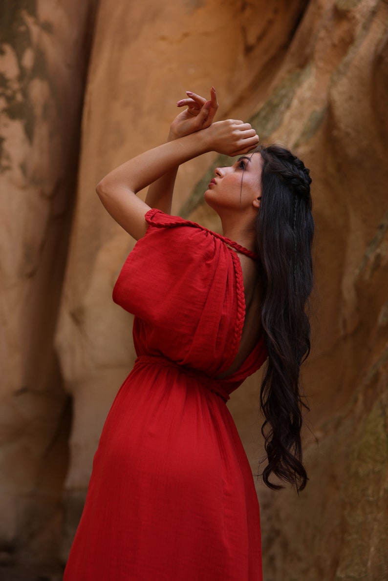 Puede incluir: Una mujer con un vestido rojo y el pelo trenzado se encuentra de pie frente a una pared rocosa. Est&aacute; mirando hacia arriba y sus manos est&aacute;n levantadas por encima de su cabeza.