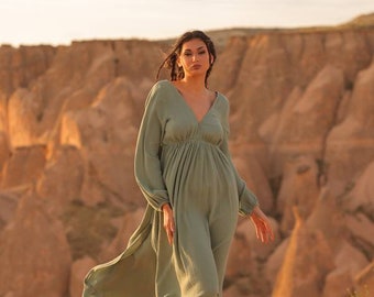 May include: A woman wearing a sage green maxi dress with long sleeves and a V-neckline stands in a desert landscape. She is wearing sandals and has her hair pulled back.
