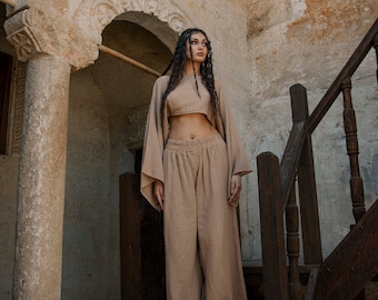 May include: A woman wearing a light brown crop top and wide-leg pants stands on a wooden staircase in a stone building.