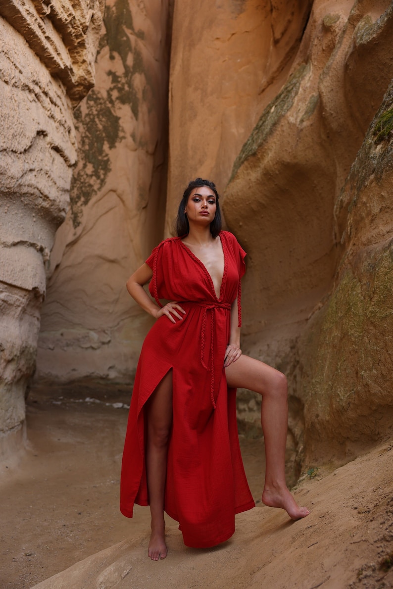 Puede incluir: Una mujer con un vestido maxi rojo con un escote en V profundo y una abertura lateral se encuentra en un ca&ntilde;&oacute;n rocoso. El vestido tiene un cintur&oacute;n trenzado y mangas cortas.