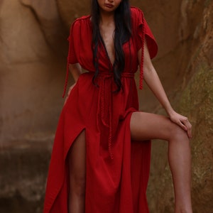 Puede incluir: Una mujer con un vestido maxi rojo con escote pronunciado y aberturas laterales. El vestido tiene un detalle de cuerda trenzada en el escote y la cintura.