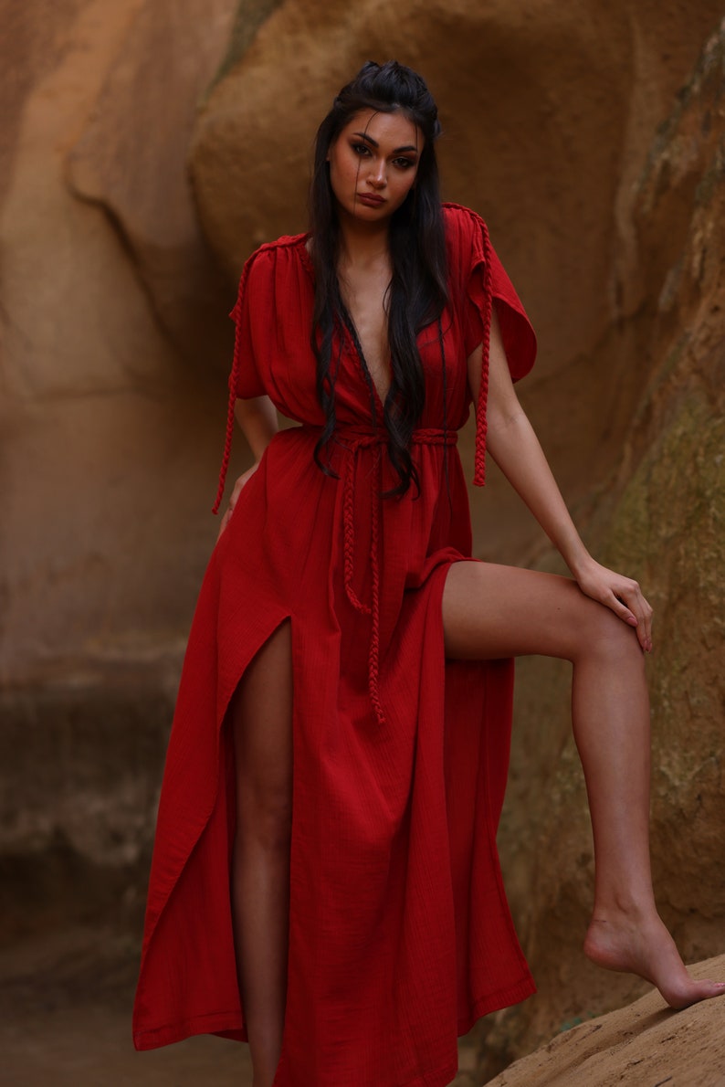 Puede incluir: Una mujer con un vestido maxi rojo con escote pronunciado y aberturas laterales. El vestido tiene un detalle de cuerda trenzada en el escote y la cintura.