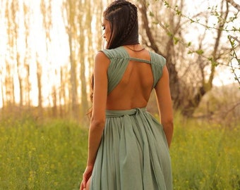 Puede incluir: Una mujer lleva un vestido maxi verde salvia con la espalda baja y una abertura en el muslo. El vestido tiene un lazo de estilo envolvente en la espalda y está hecho de una tela fluida. Está de pie en un campo de hierba verde con árboles en el fondo.