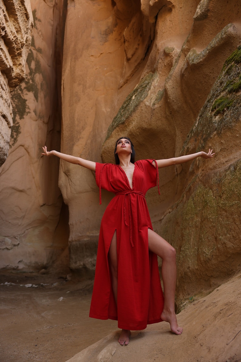 Puede incluir: Una mujer con un vestido maxi rojo con cuello en V profundo y aberturas laterales se para con los brazos extendidos en un ca&ntilde;&oacute;n rocoso.
