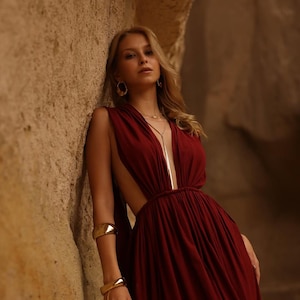 Puede incluir: Un vestido sin mangas de color burdeos intenso con un escote en V pronunciado, diseño abierto en los laterales y cintura fruncida. La prenda se complementa con pendientes de aro dorados, un collar dorado de varias capas y dos brazaletes.