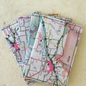 Może przedstawiać: Stos złożonych map przedstawiających różne obszary Montany, w tym Great Falls, Billings i Hardin. Mapy są wydrukowane w czerni i bieli z czerwonymi i niebieskimi akcentami. Mapy są złożone i ułożone na jasnobeżowej powierzchni.